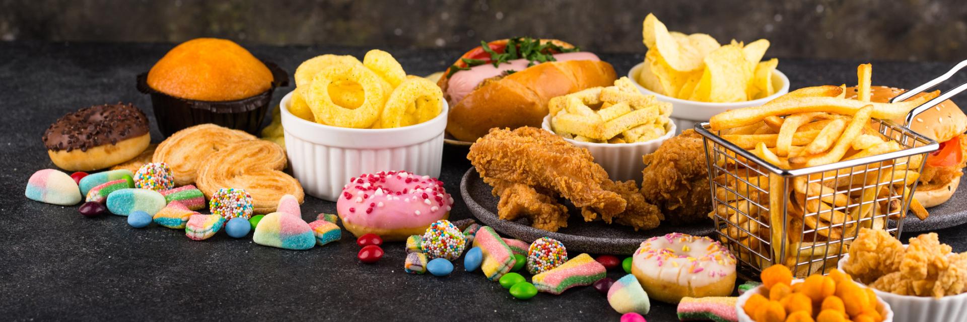 An assortment of ultra-processed foods including donuts/pasties, candy, and fried foods such as potato chip, onion rings, fries, and breaded chicken.