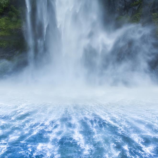 Shot from the top of the waterfall Milford Sound in New Zealand. The waterfall creates symmetrical patterns, and there's moss vegetation in the background. 